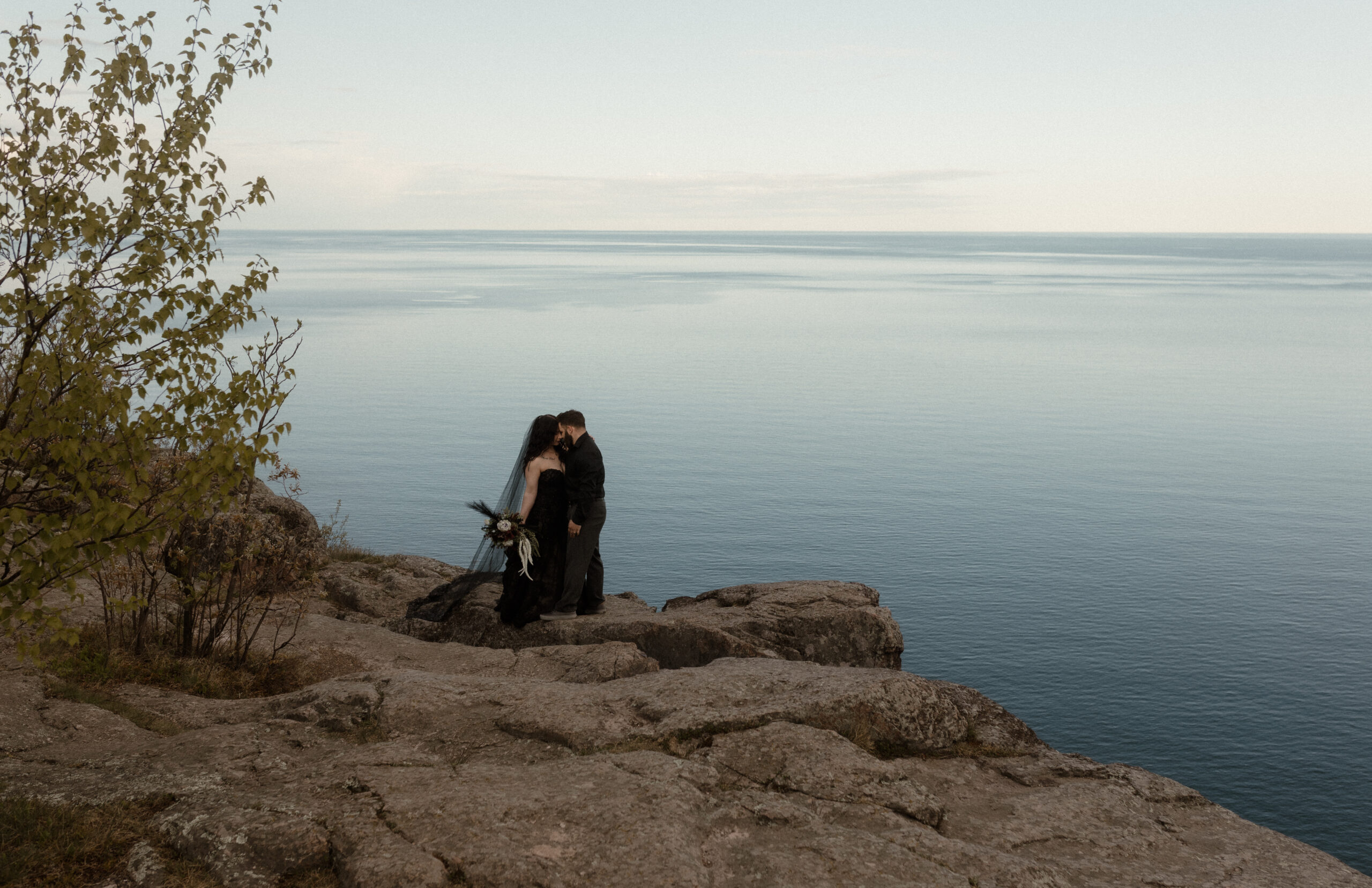 A couple dressed in all black stands on a rocky cliffside overlooking the still blue waters of Lake Superior. The bride wears a black gown with a flowing black veil and holds a moody bouquet, while the groom gently leans in to kiss her. The serene lake stretches to the horizon under a soft, pale sky, with early green leaves framing the left side of the image.